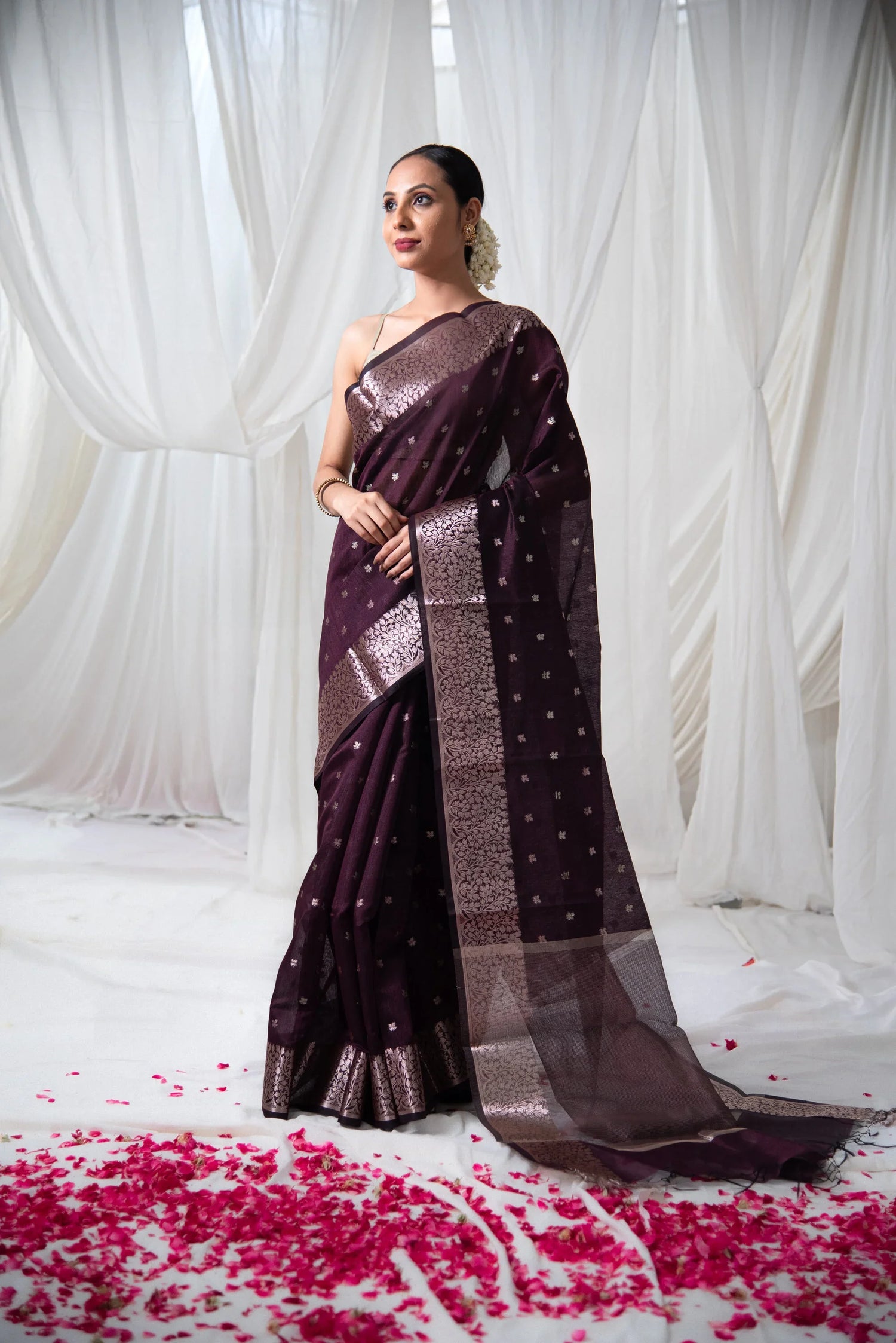 Woman in maroon Banarasi cotton saree with gold detailing, standing on rose petals