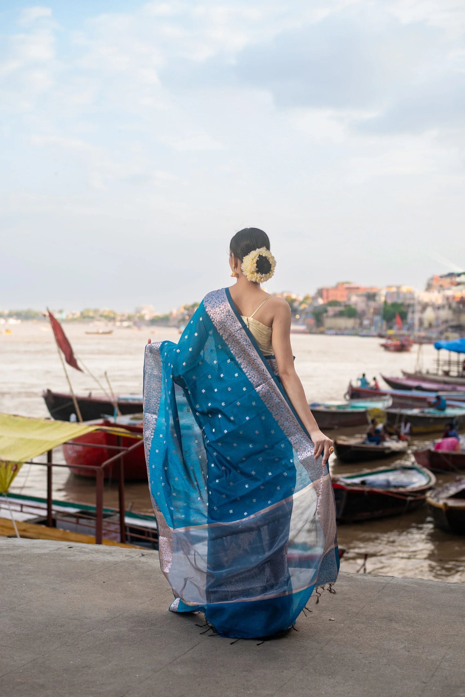 Woman in blue Banarasi saree with zari work by riverside, boats in background