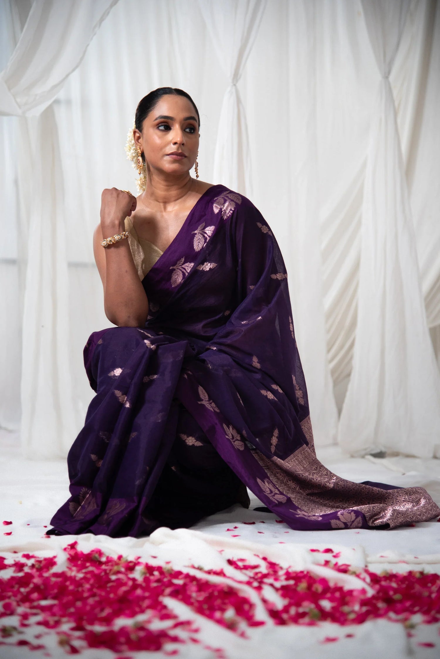 Woman wearing a purple Banarasi cotton saree with gold floral motifs, seated indoors with rose petals on the floor.