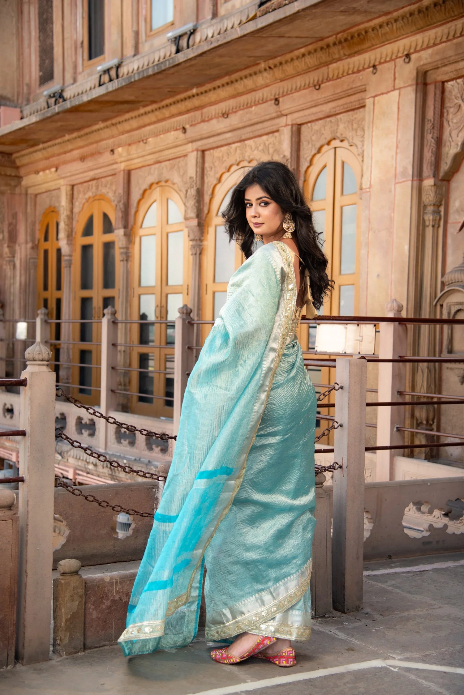 Woman in light blue Banarasi silk saree with gold border standing on a heritage balcony