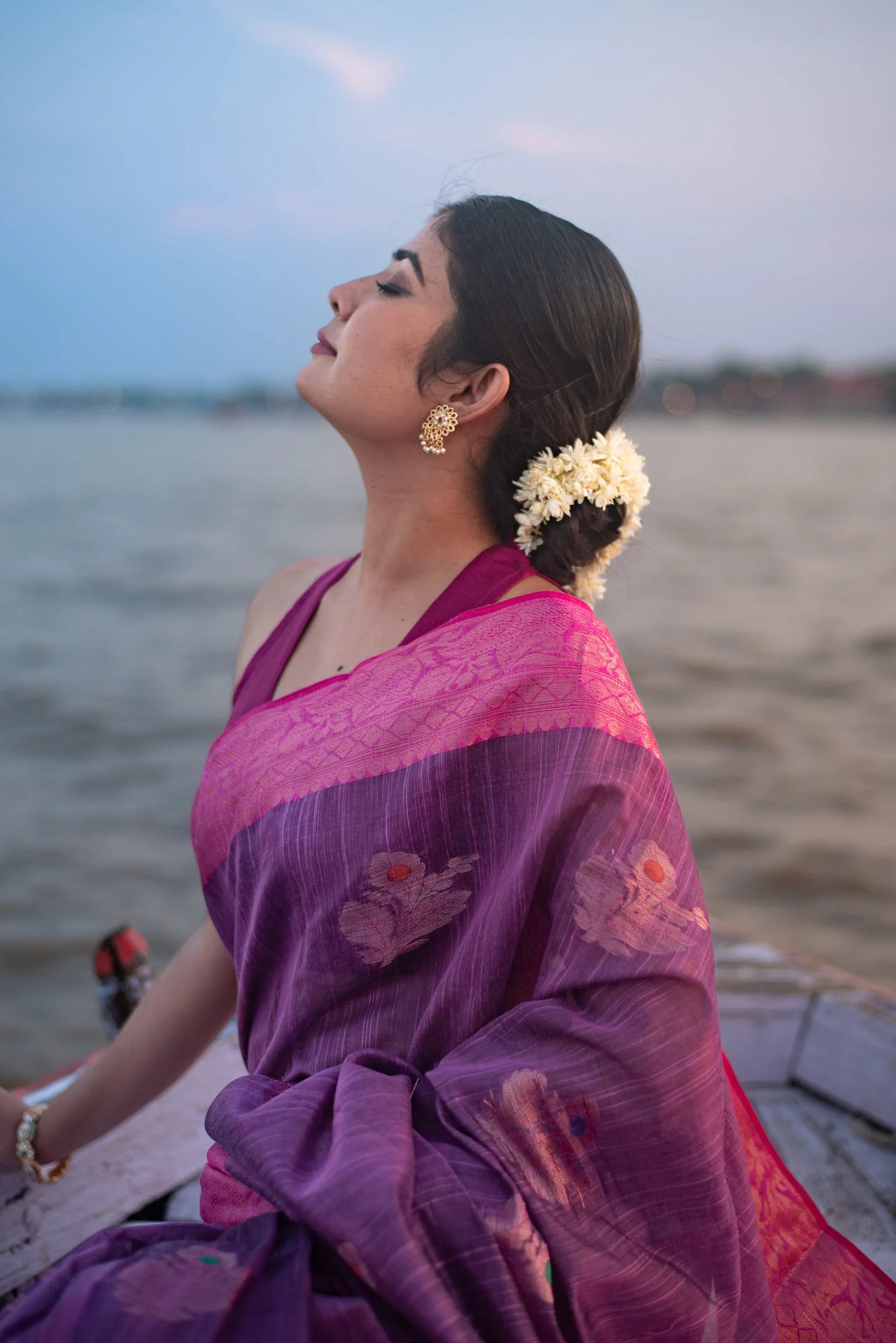 Woman by river in magenta dual-tone Banarasi silk saree with floral motifs, elegant bun with flowers