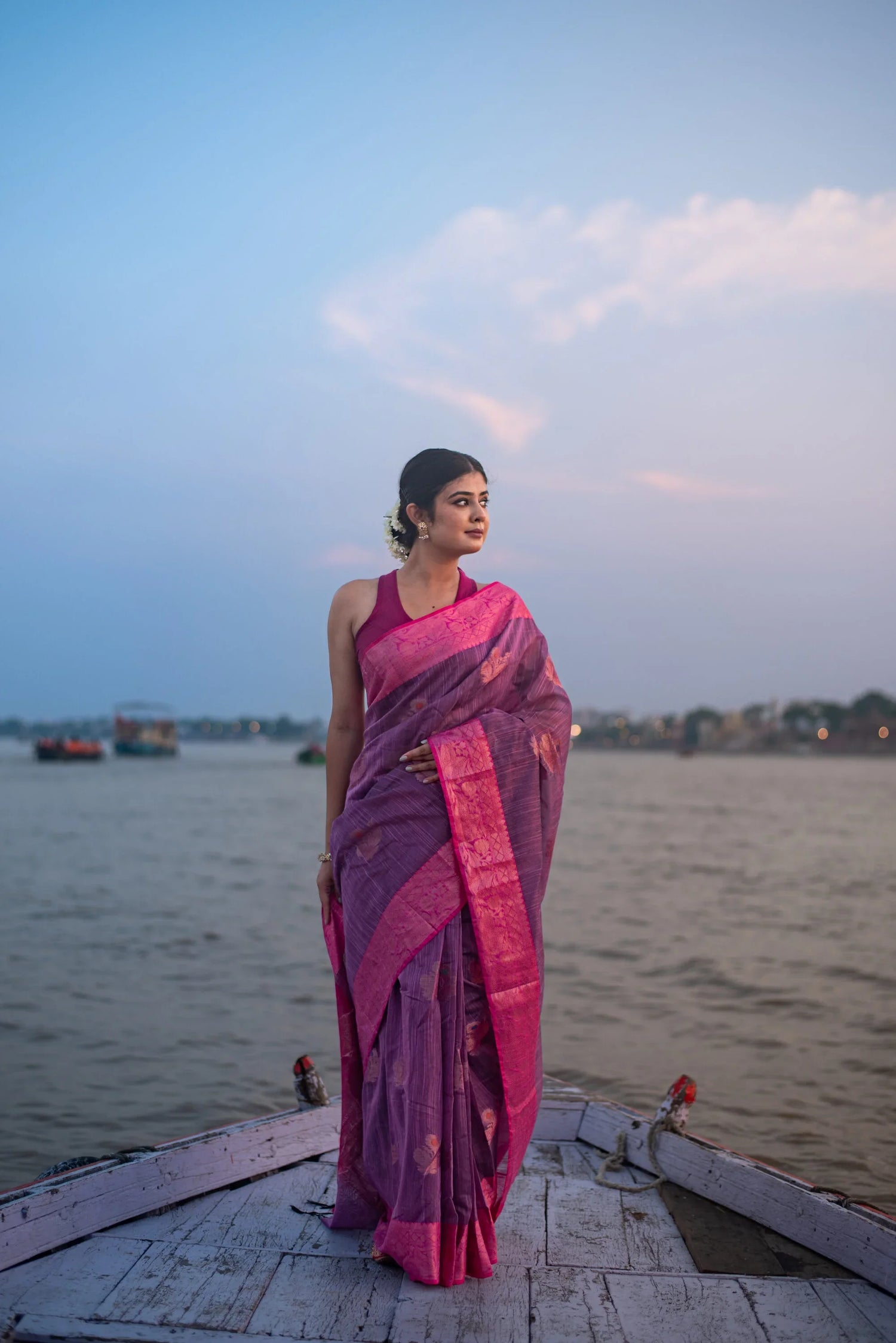 Woman in a purple and pink Banarasi silk saree standing on a boat by the river at sunset