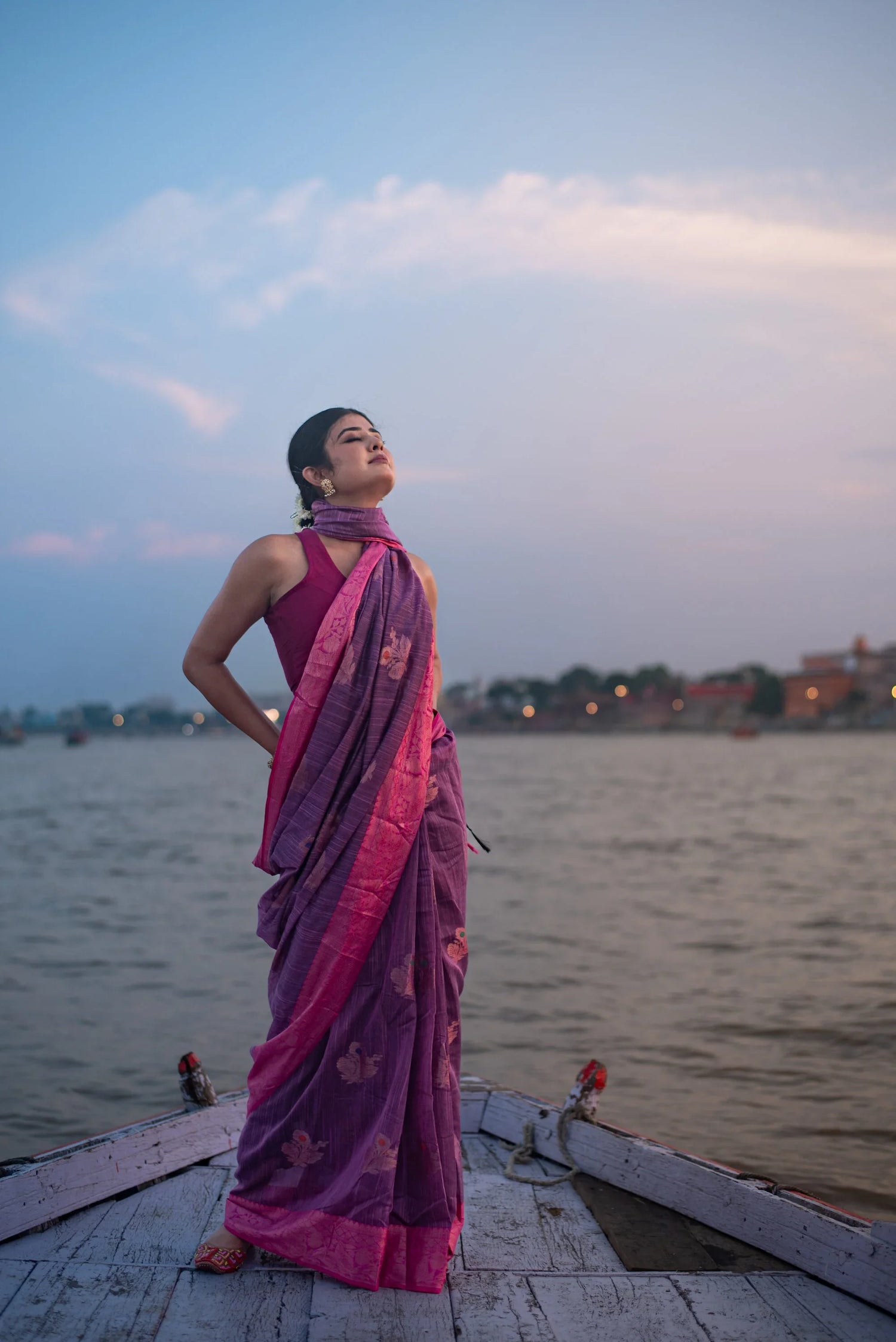 Woman in a purple and pink Banarasi silk saree standing on a boat by the river at sunset