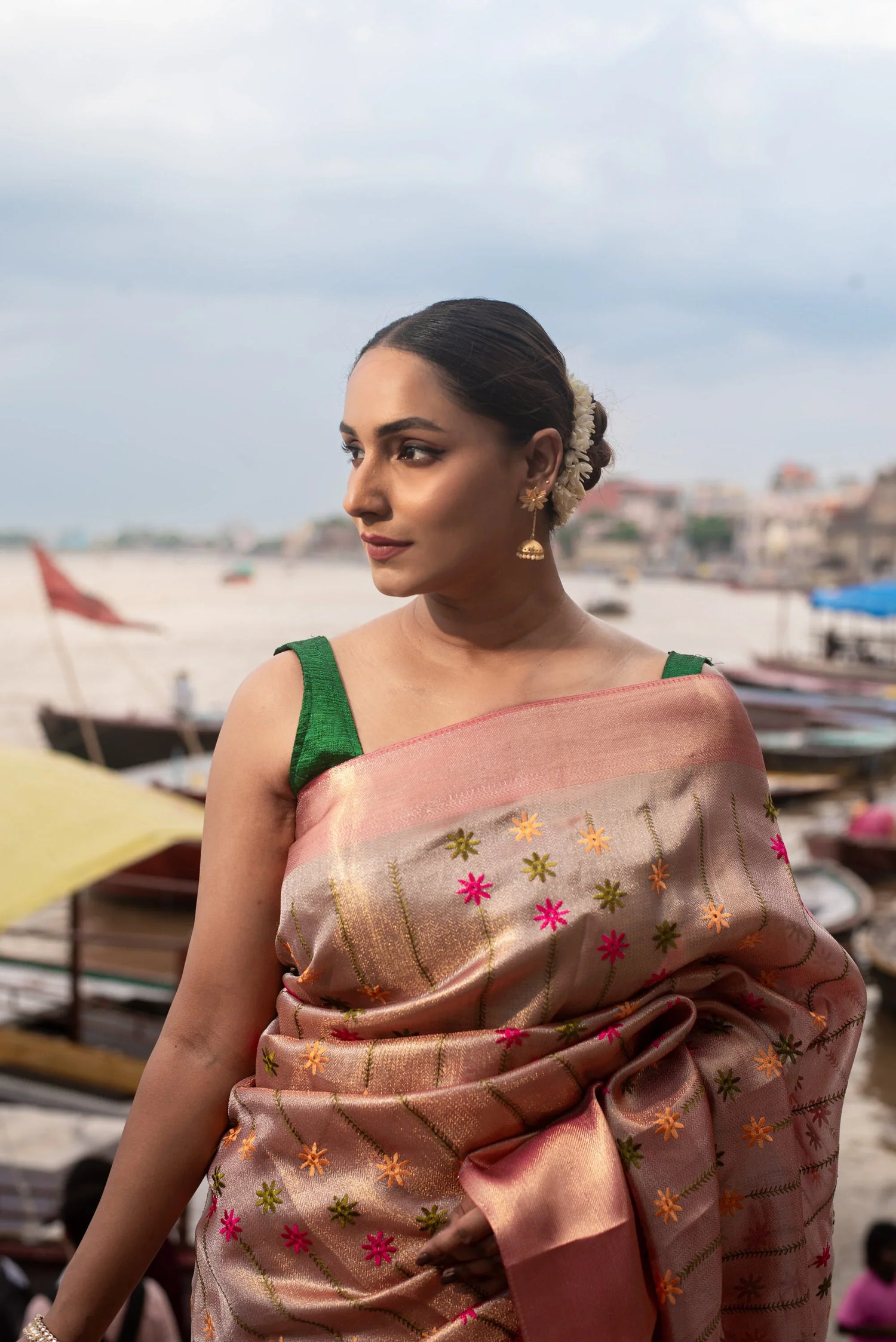 Woman wearing pink Banarasi silk saree with floral meenakari near riverside ghats
