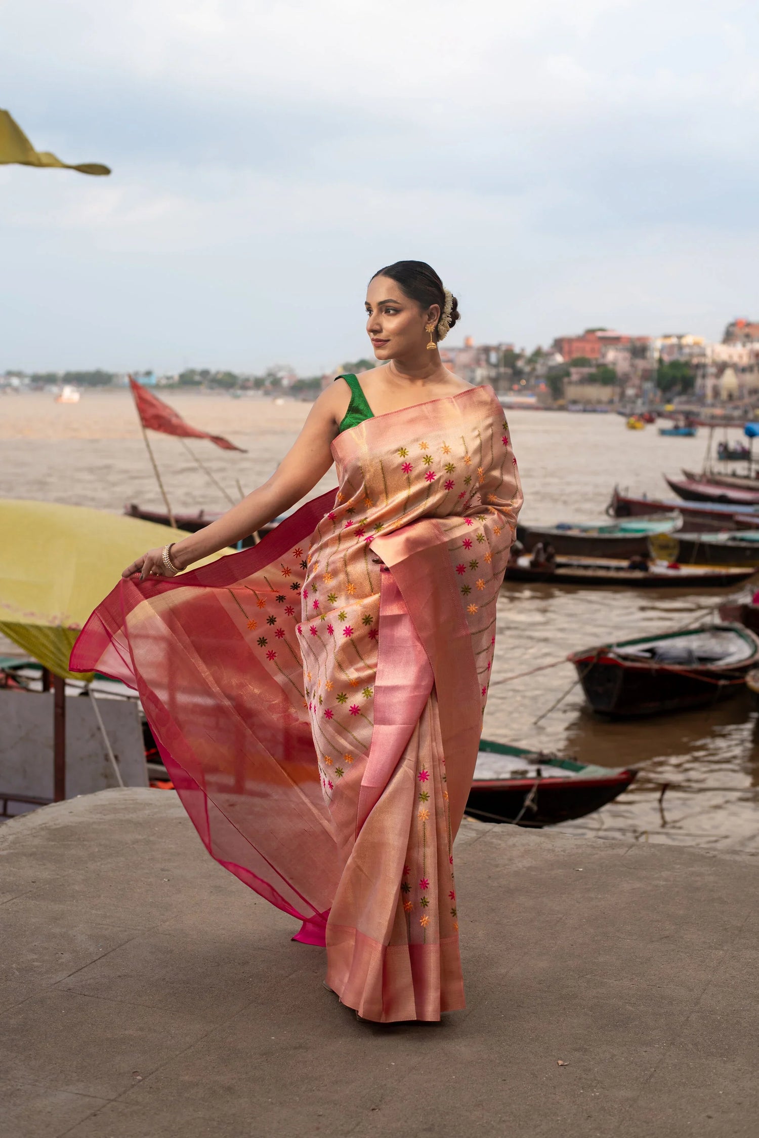 Woman wearing pink tissue Banarasi saree with Meenakari work by riverside with boats