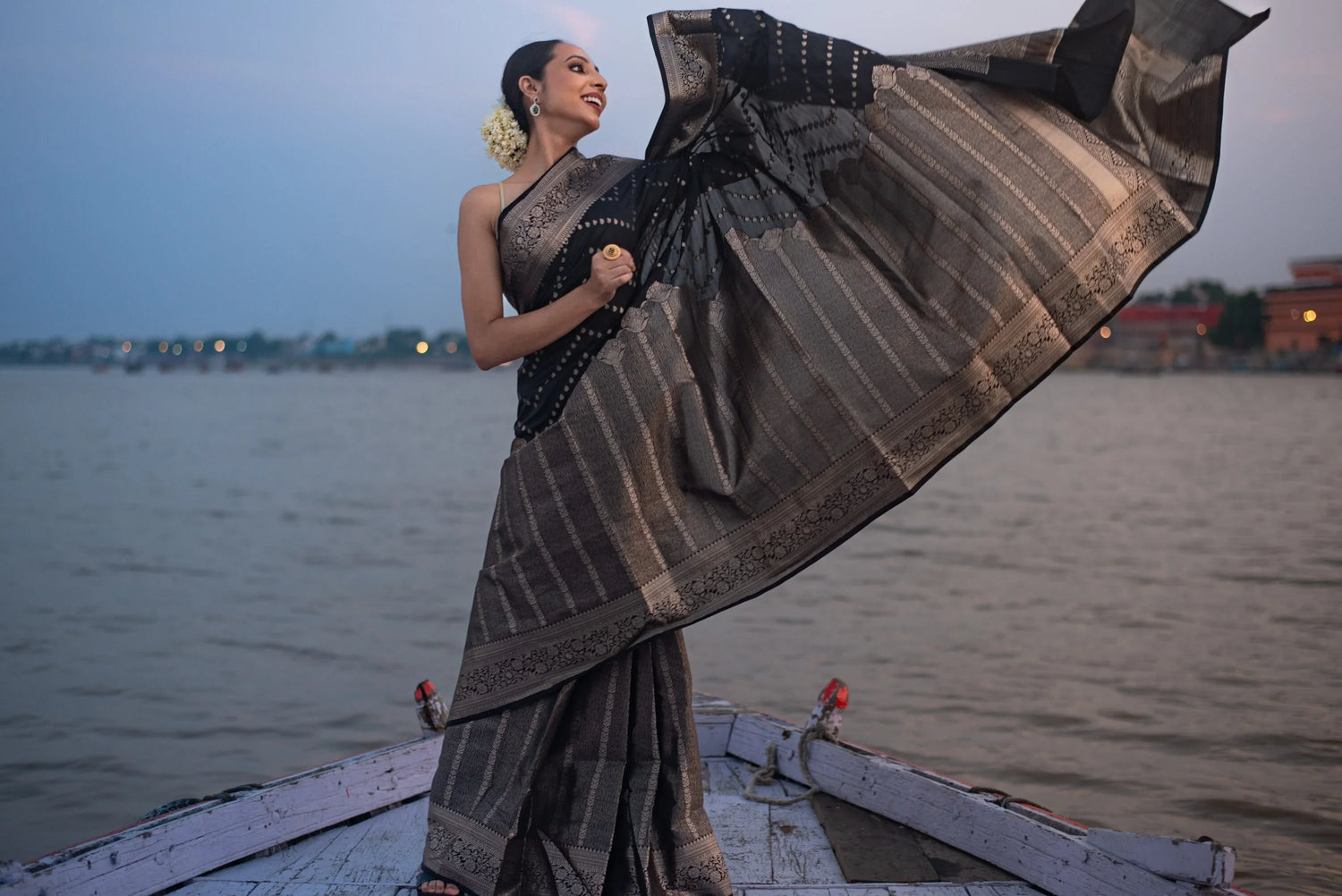 Woman wearing a black Banarasi Katan silk saree with zari work, standing on a boat by a river.