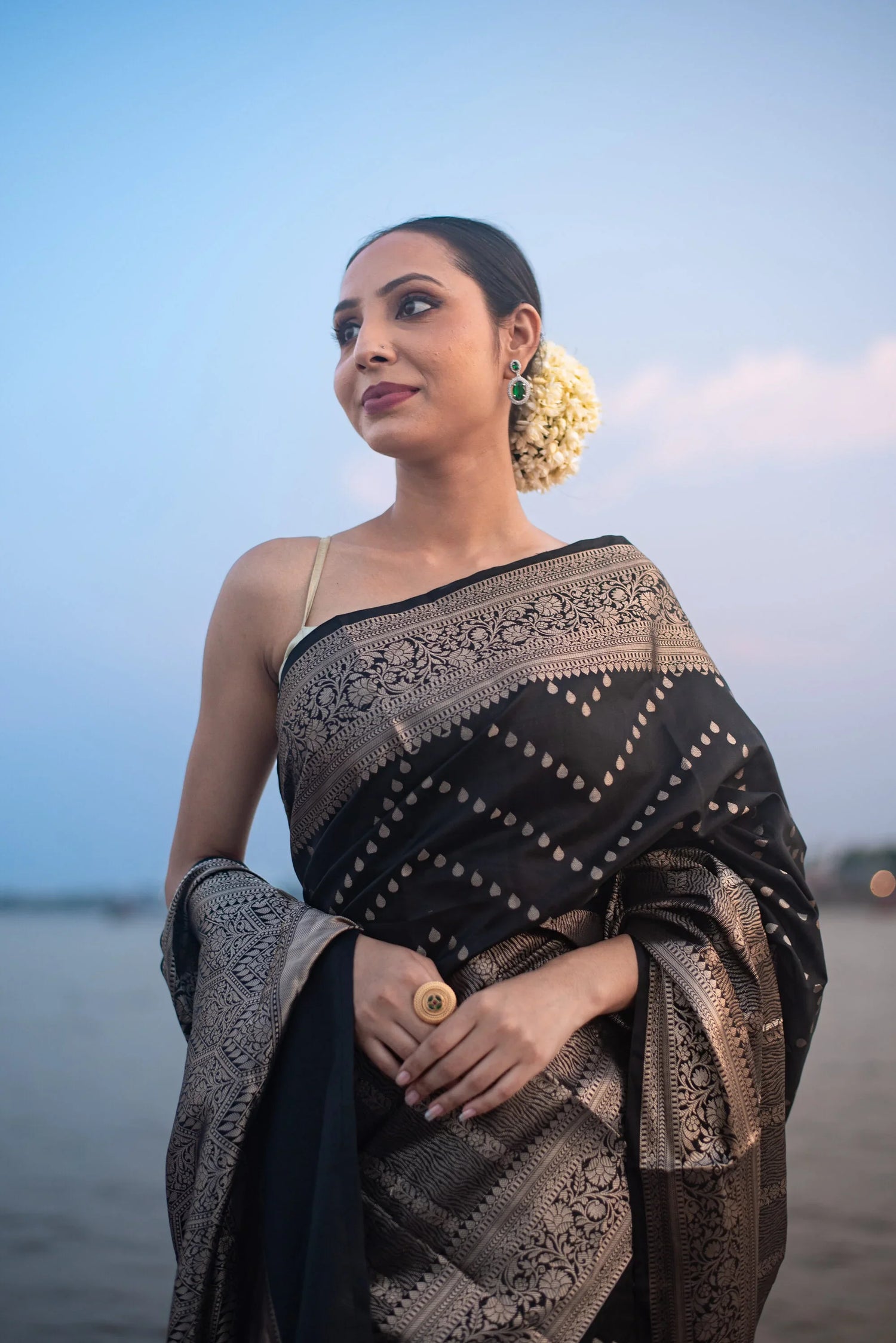 Woman in a black Banarasi Katan silk saree with intricate zari work by the water at dusk.
