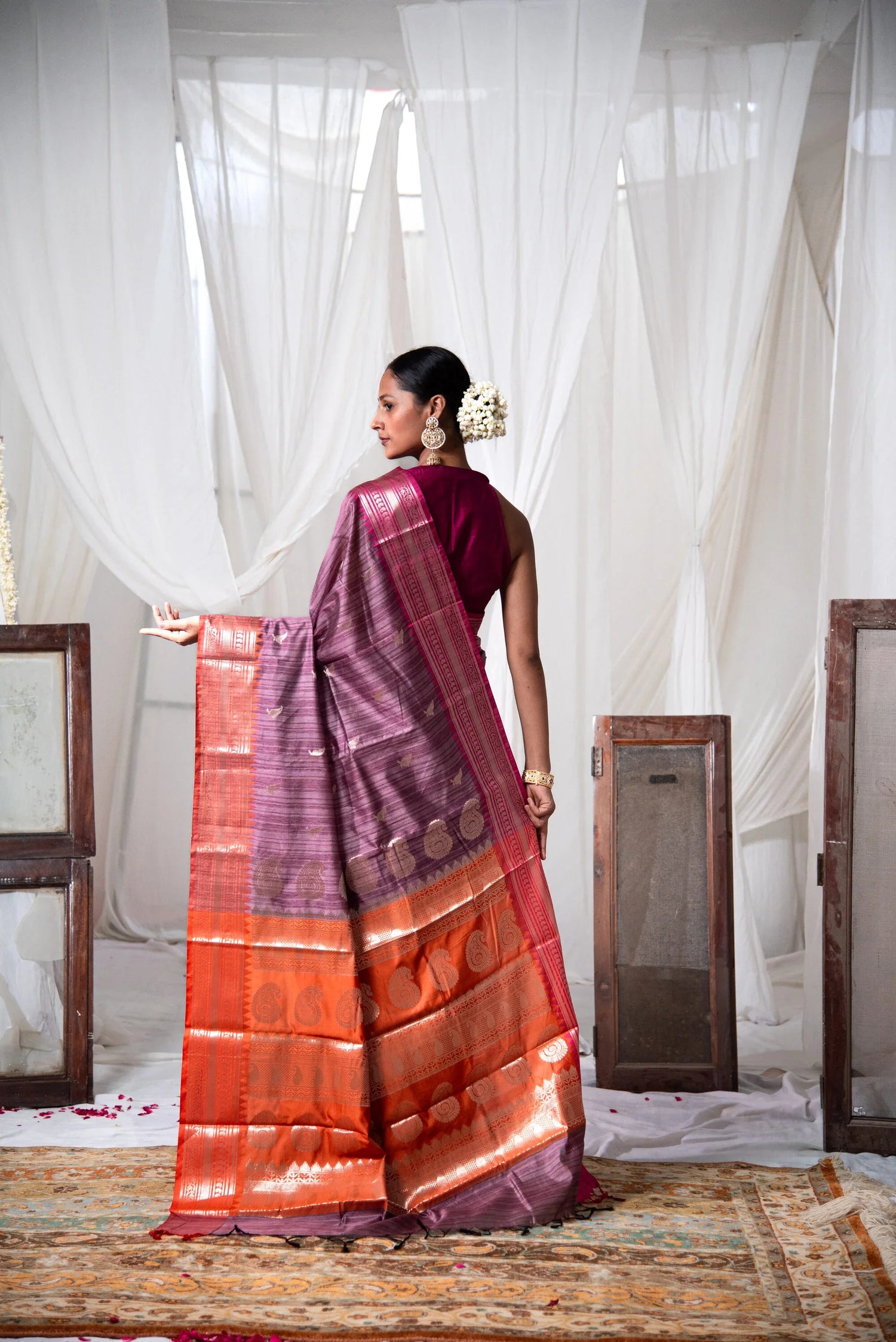 Woman in a dual-tone orange and purple cotton saree with zari work, standing in a traditional setting