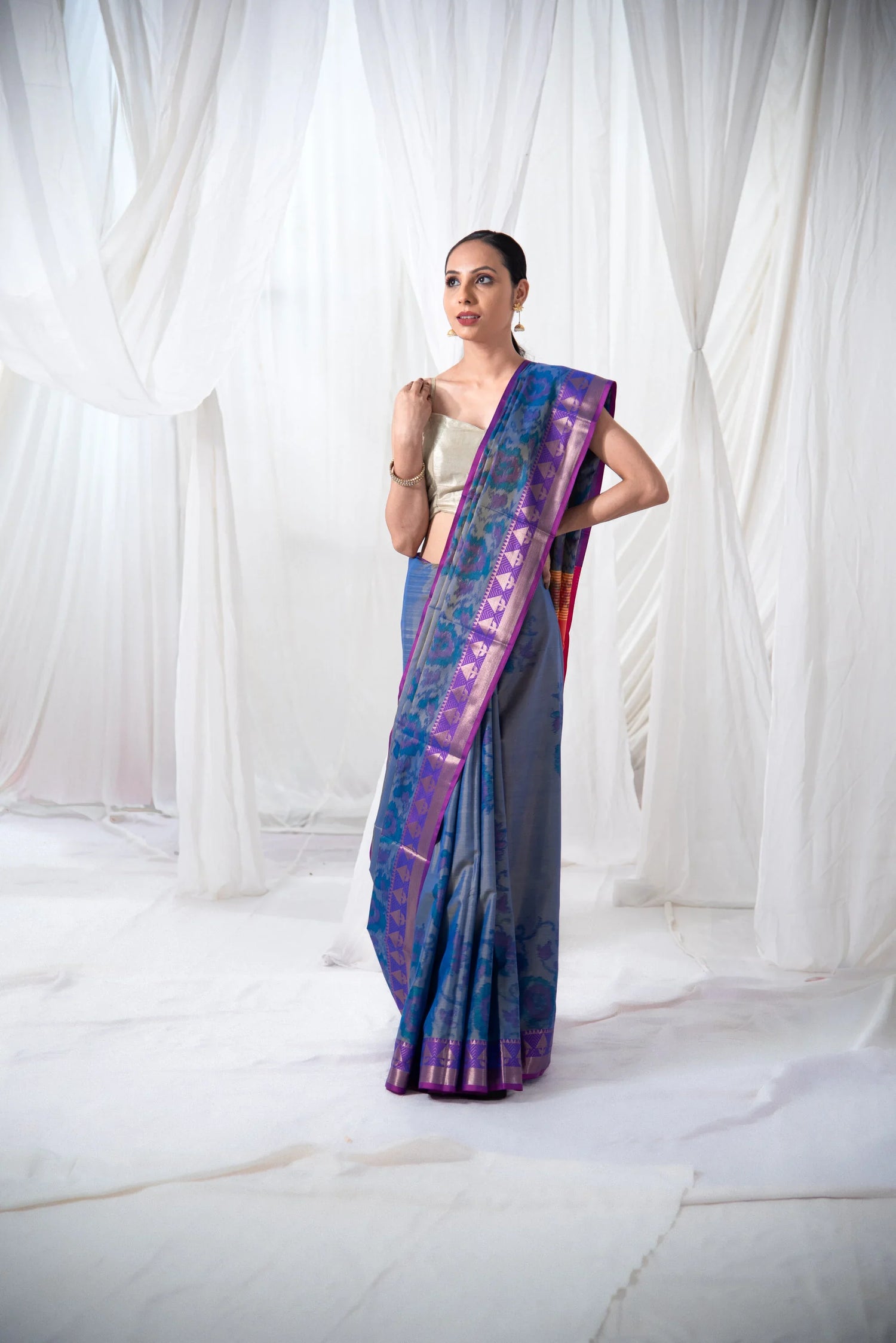 Woman wearing a blue Ikkat Banarasi silk saree with purple border, standing indoors.