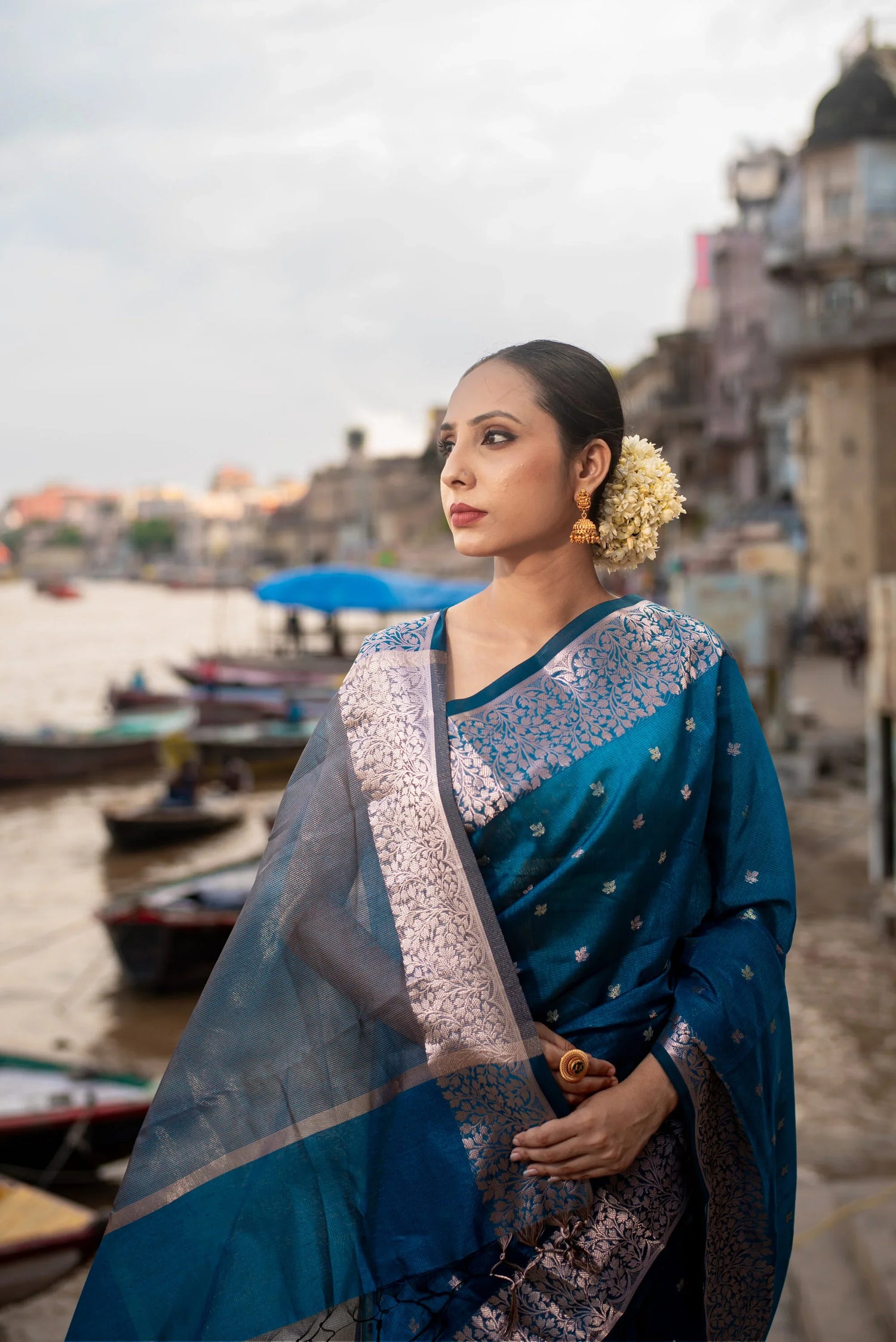 Woman in blue Banarasi cotton saree with zari work, standing by riverside, floral gajra in hair