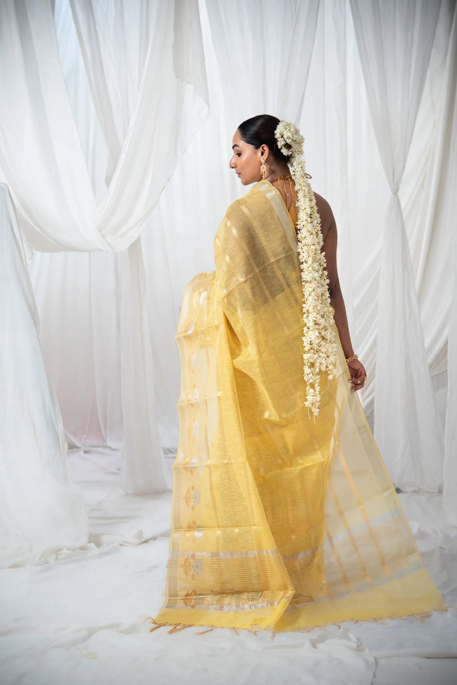 Woman in yellow Banarasi silk saree with floral hair garland, white draped backdrop