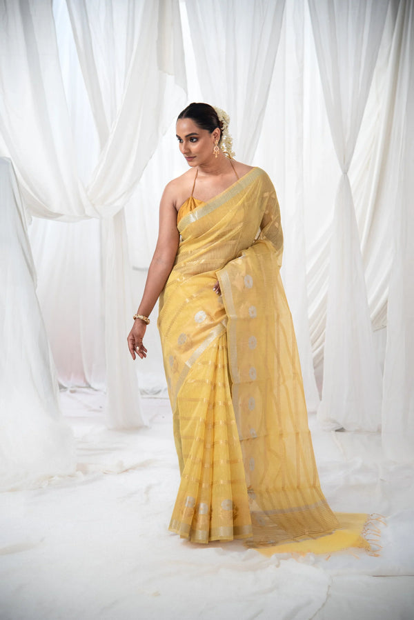 Woman in yellow Banarasi cotton linen silk saree with silver zari, floral motifs, in white draped setting — alternate view (back / drape detail)