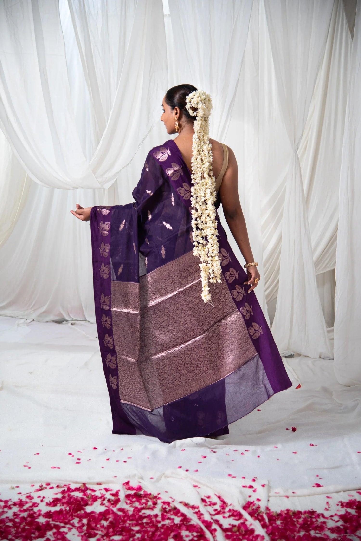Woman in purple Banarasi cotton saree with gold floral motifs, floral hair garland, backdrop of white drapes