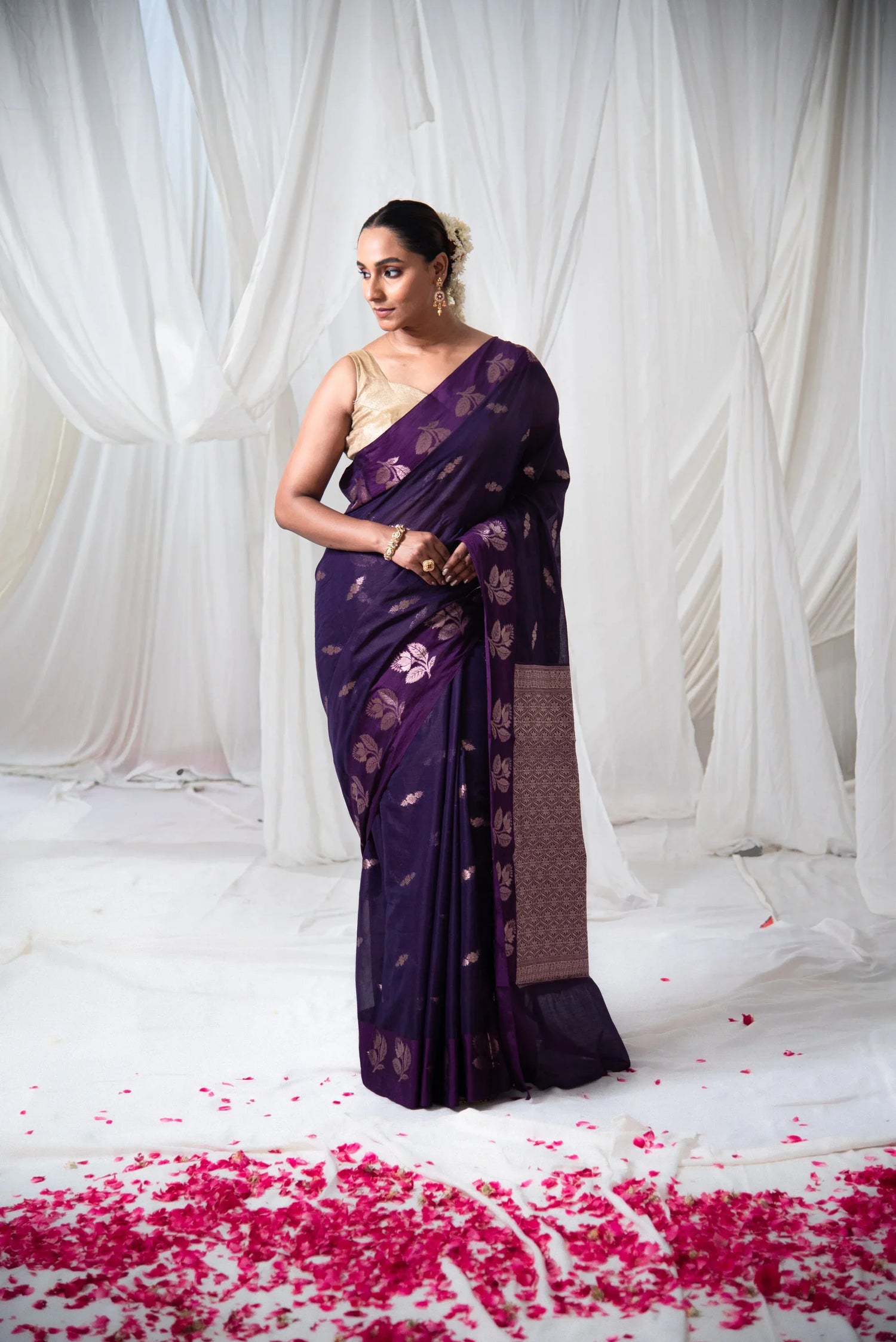 Woman in a purple Banarasi cotton saree with gold motifs, standing on white fabric with rose petals