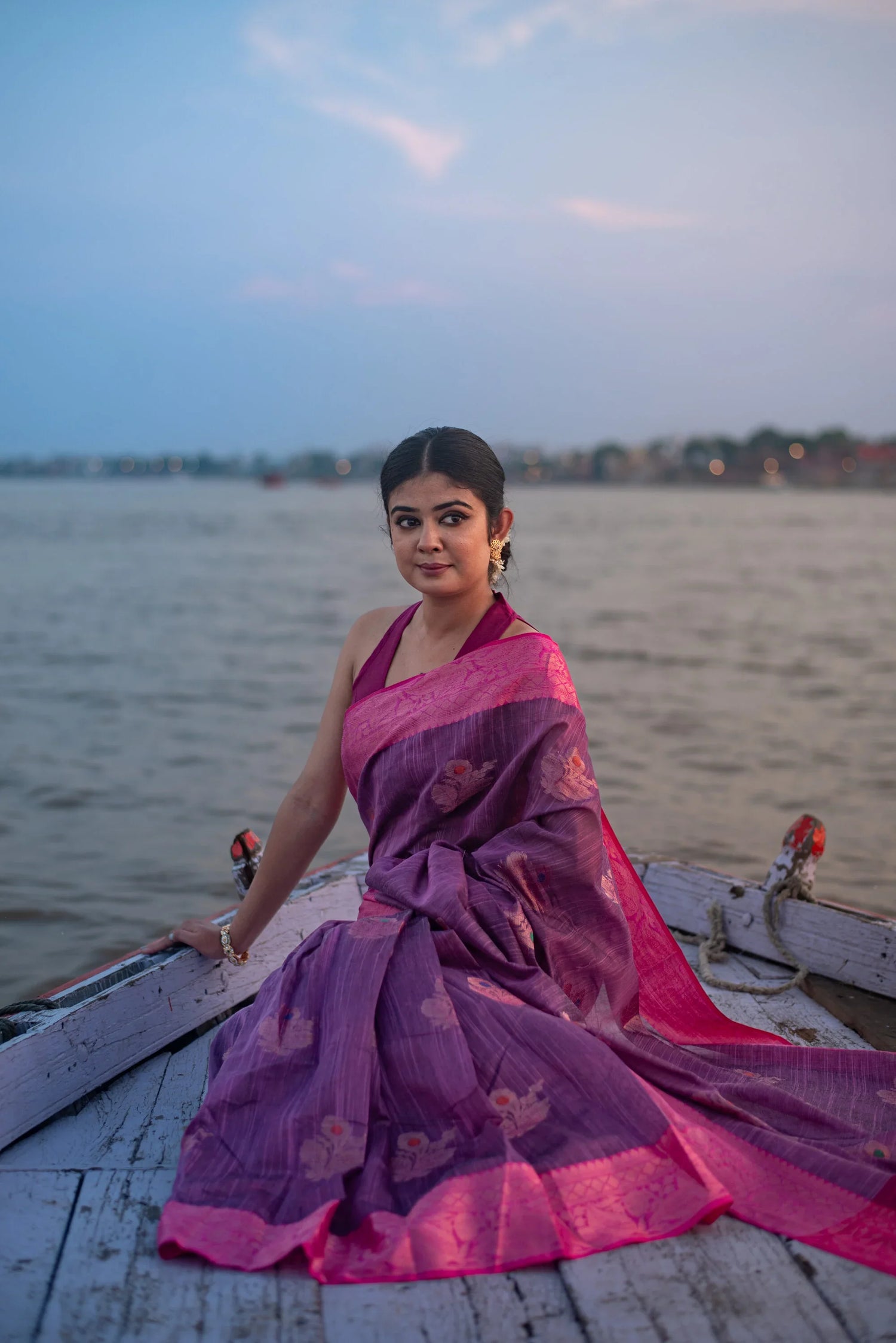 Woman in purple dual-tone Banarasi silk saree sitting on a boat by the riverside at dusk