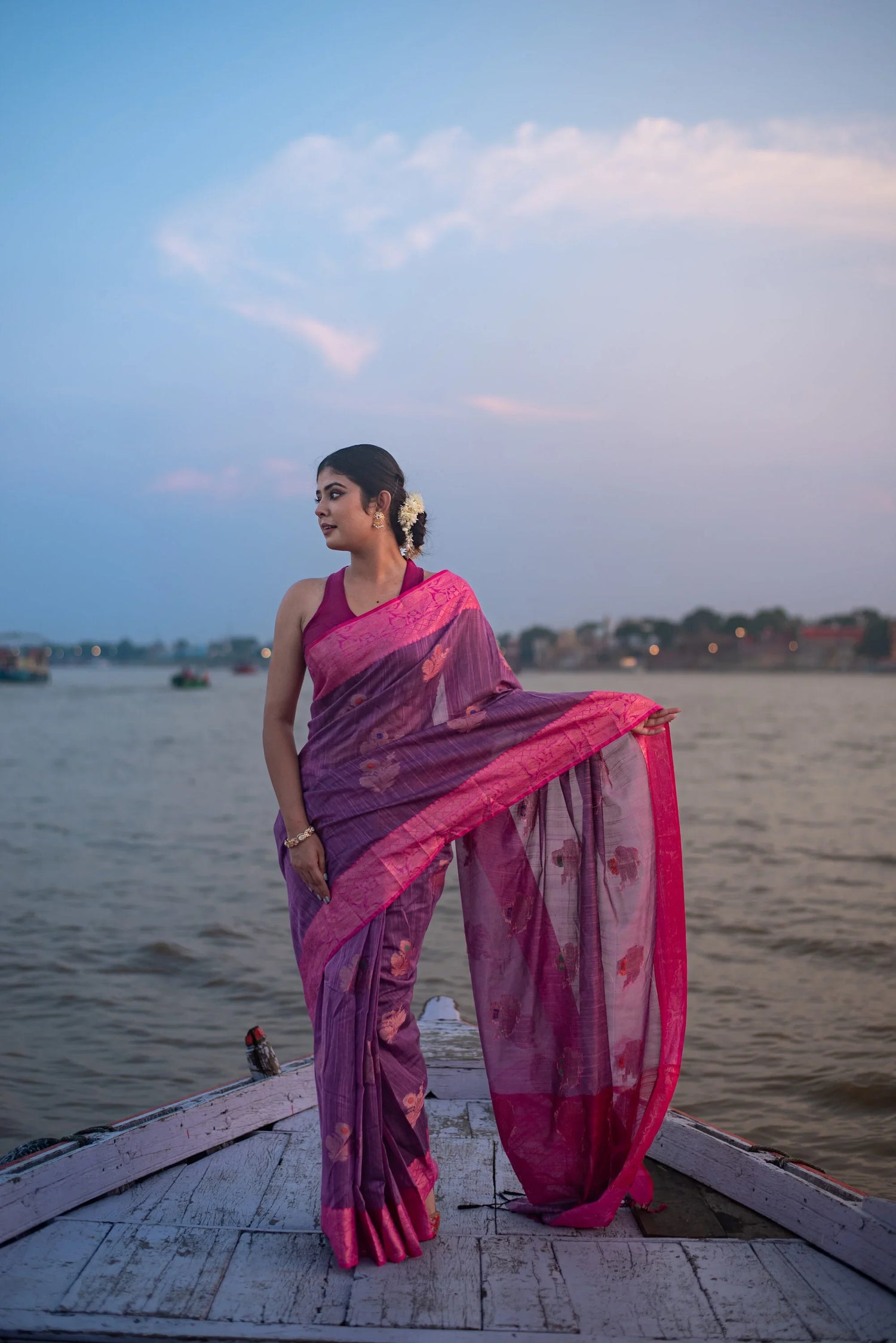 Woman in purple and pink Banarasi silk saree standing on a boat by the river at dusk