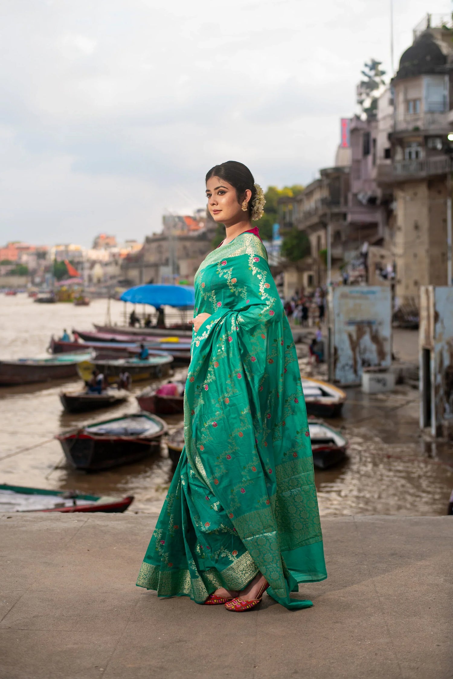 Woman in green Banarasi silk saree with Meenakari work by riverside ghats, boats in background
