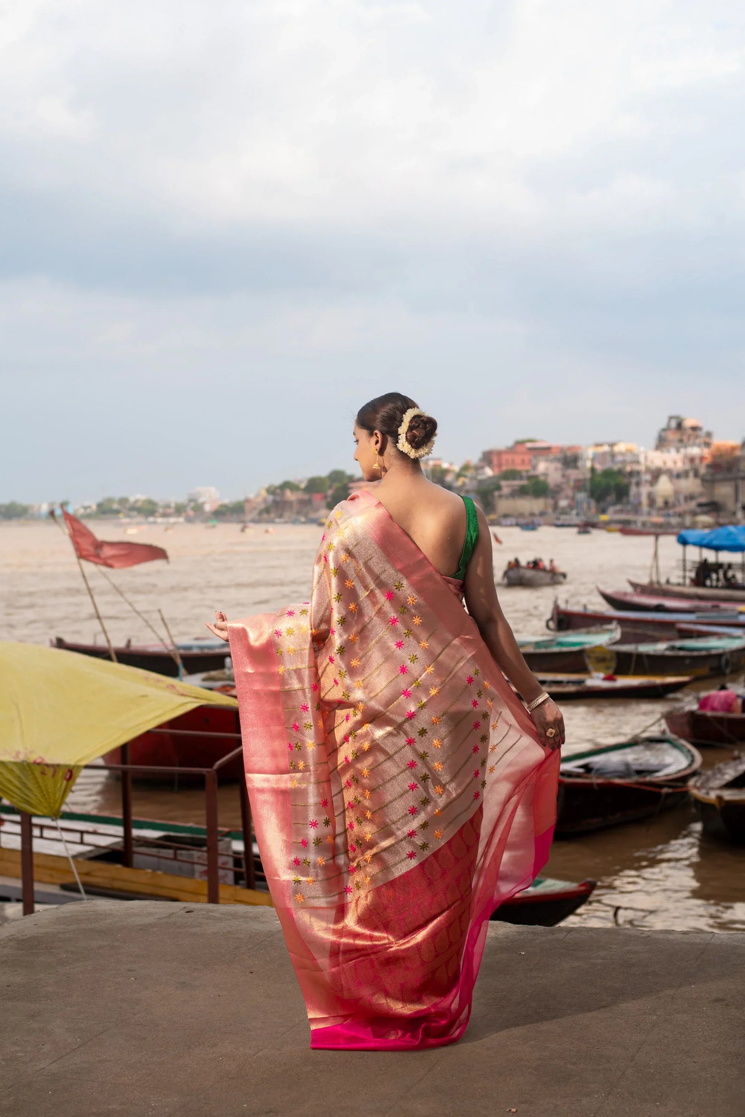 Woman in pink Banarasi Meenakari tissue saree by riverside with boats in background