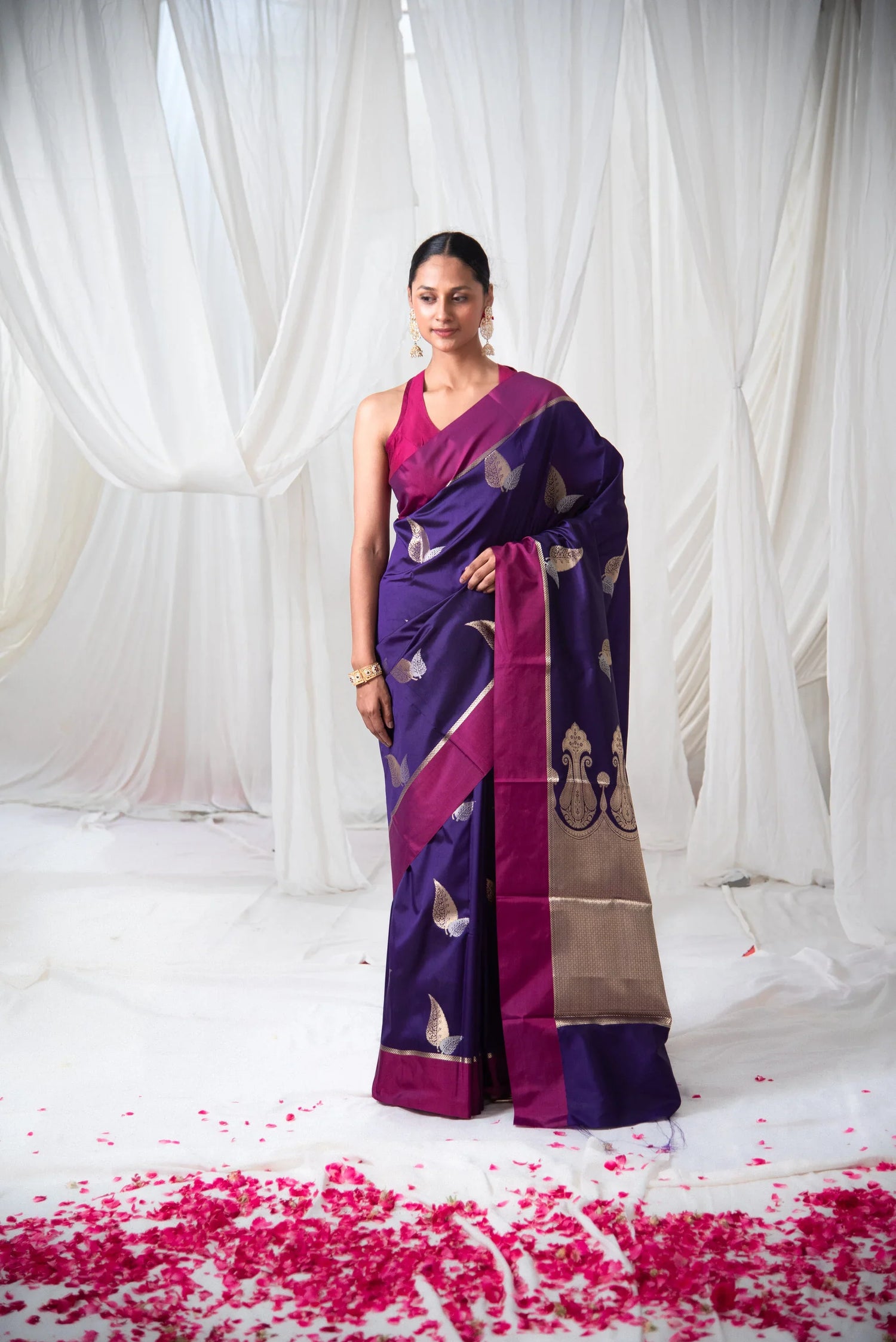 Woman wearing a purple Banarasi silk saree with gold motifs and magenta border, floral backdrop