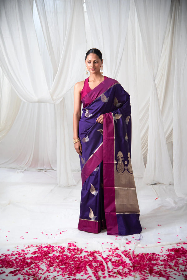 Woman wearing a purple Banarasi silk saree with gold motifs and magenta border, floral backdrop
