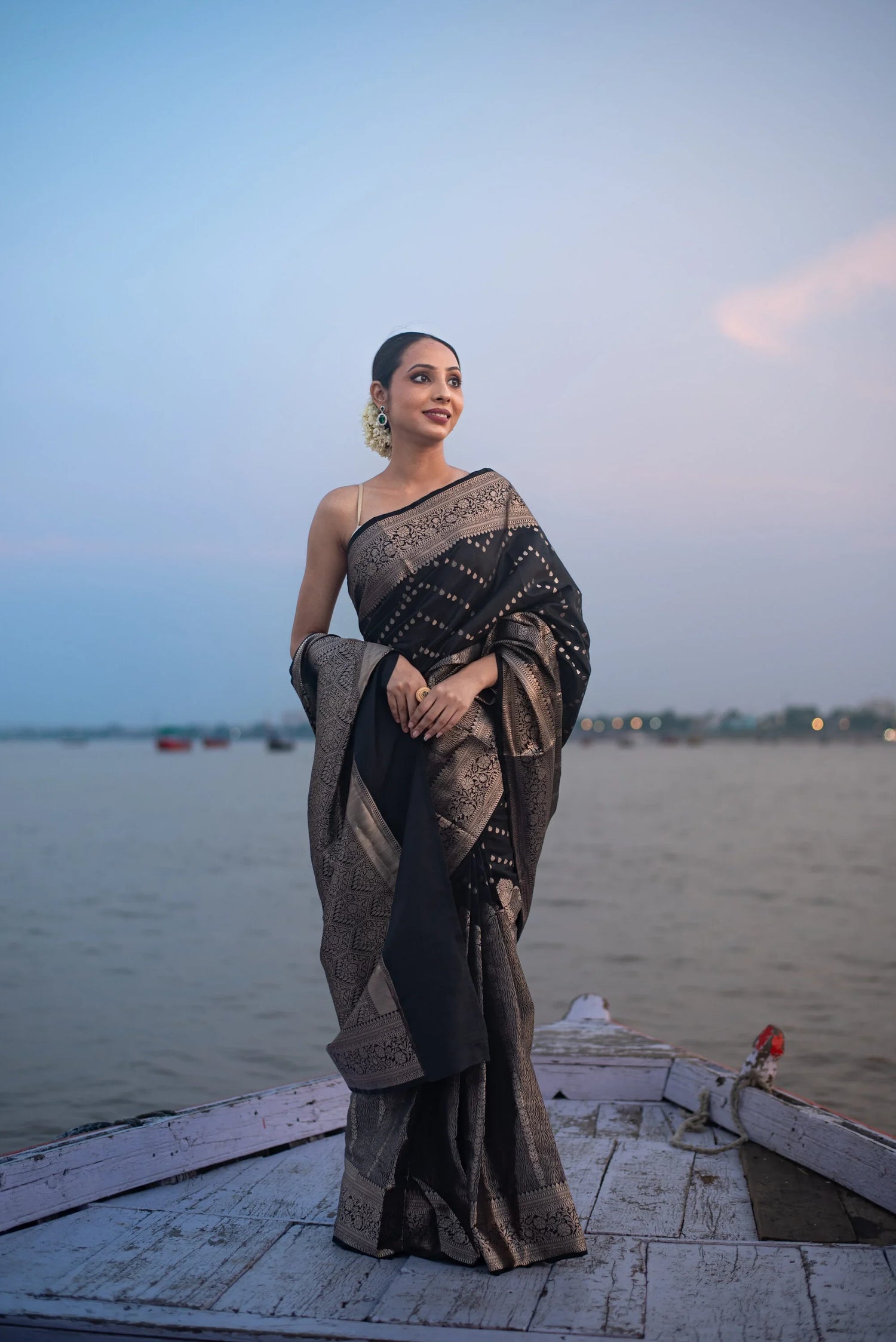 Woman in black Banarasi Katan silk saree with zari work standing by a river at sunset